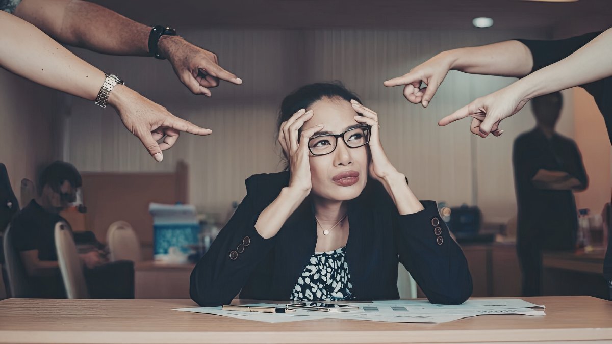 Woman sitting at a desk looking stressed while fingers point at her from different directions.